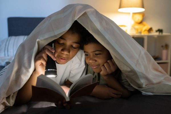 black kids lying on bed and hiding under duvet while reading book with flashlight at night