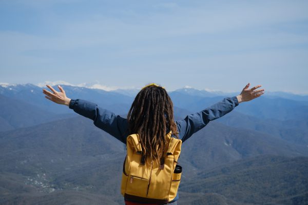 Concept travel. Caucasian woman with dreadlocks and yellow backpack admires nature of snowy peaks of Caucasian ridge. Rear view. Girl spread arms in different directions as if embracing the world.